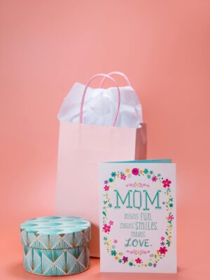 Charming Mother's Day setup with a greeting card, gift bag, and decorative box on a pastel pink background.