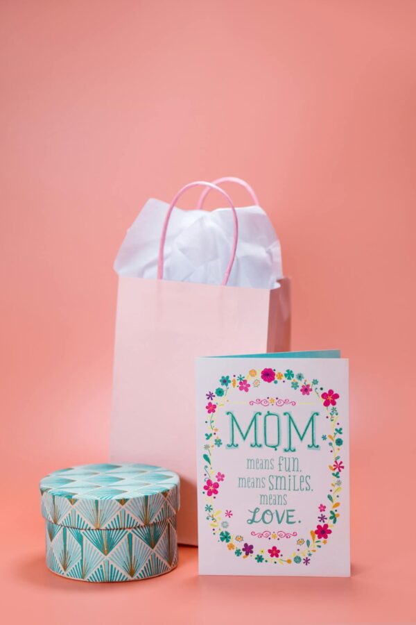 Charming Mother's Day setup with a greeting card, gift bag, and decorative box on a pastel pink background.